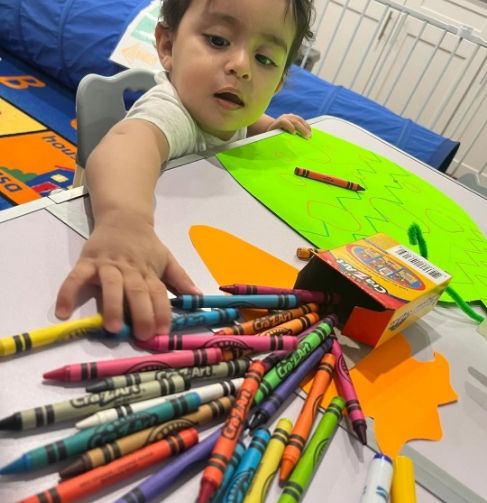 Young child at Bee Smart Preschool Education reaching for colorful crayons while drawing, developing fine motor skills and creativity.