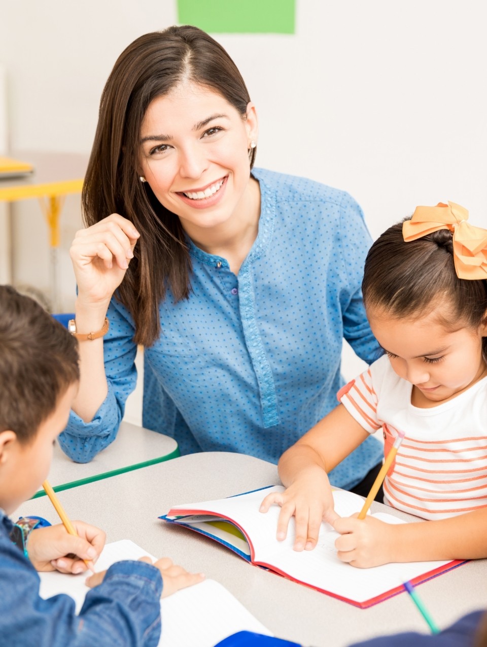 Teacher at Bee Smart Preschool Education smiling while helping young children with writing activities, promoting creativity, confidence, and early learning in a warm classroom environment.